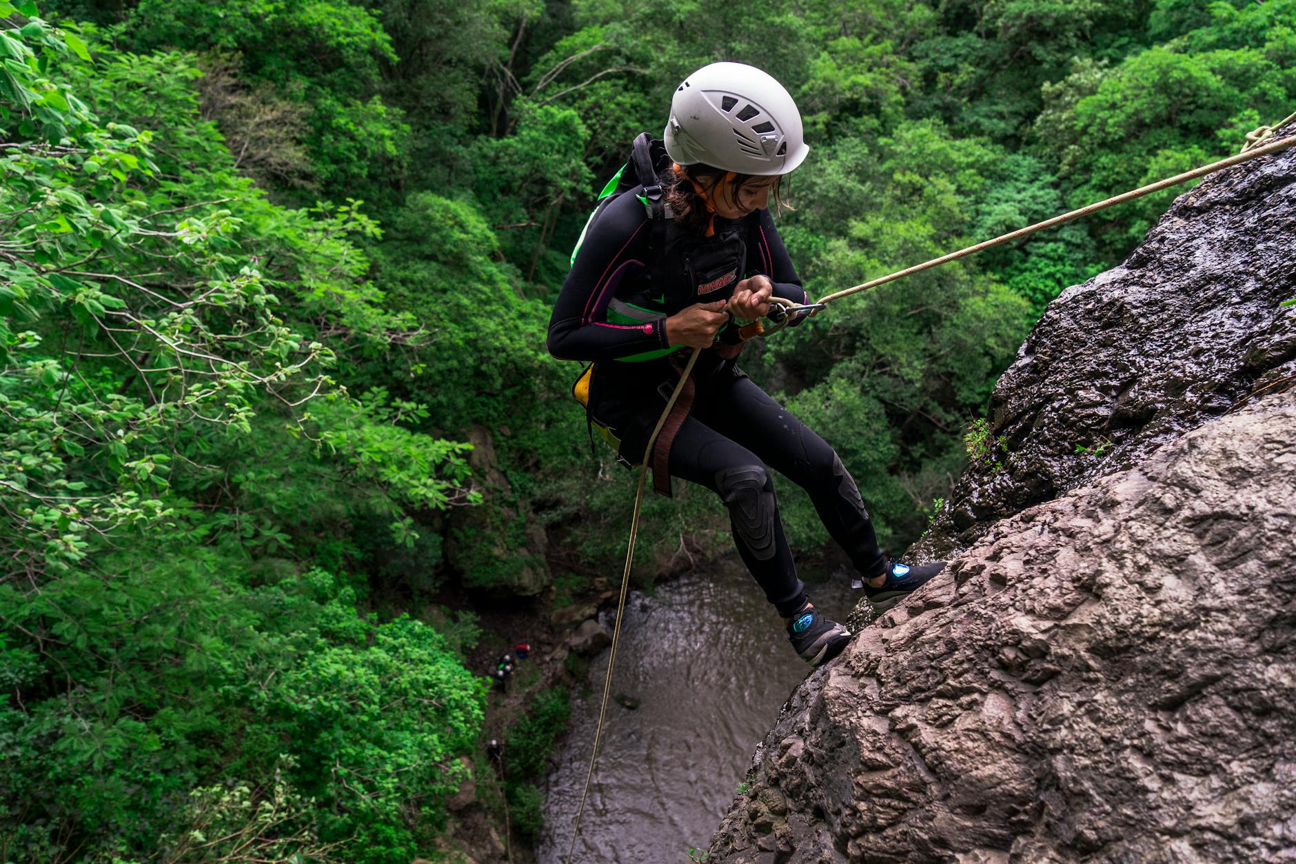 Female climber rappels down a rock cliff amidst dense green forest scenery.